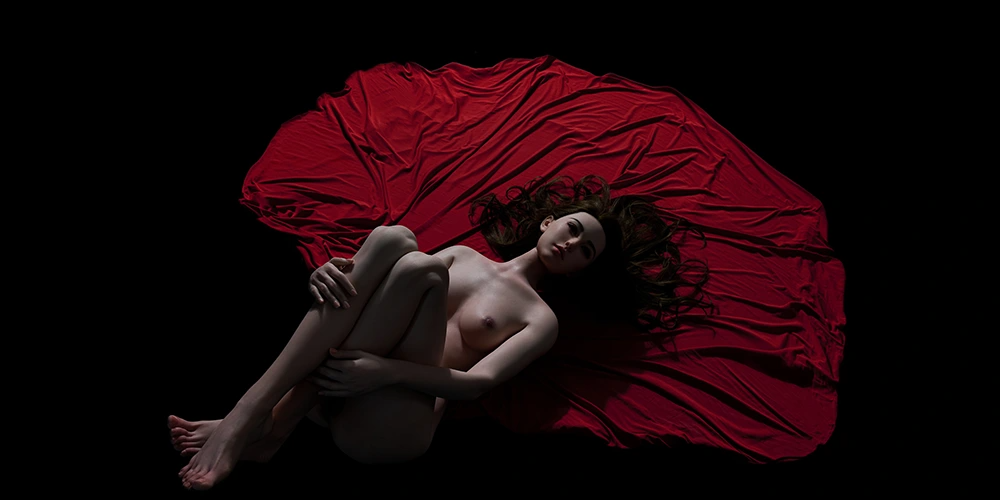 Realistic female doll lying on red fabric against a dark background.