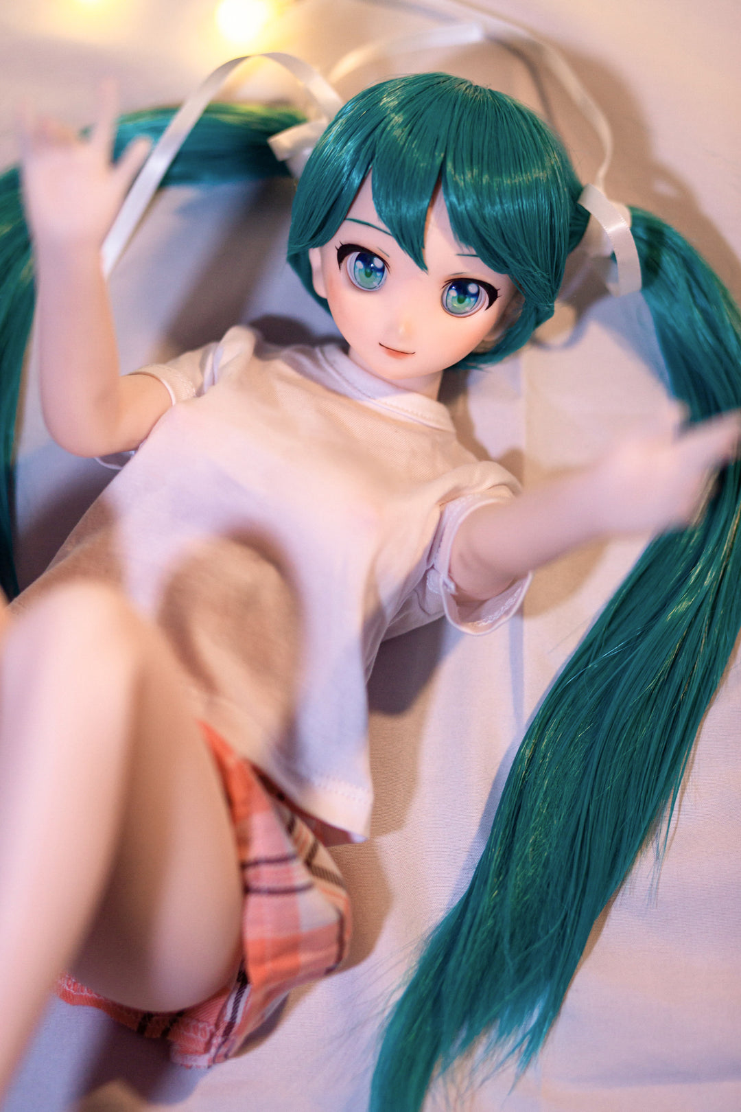 CLIMAX DOLL Mini J60 cm Hybrid - Miku (Cinnamon) - Reclining pose on white bedding with teal twin tails fanned out, white tee and plaid skirt, knees gently bent.