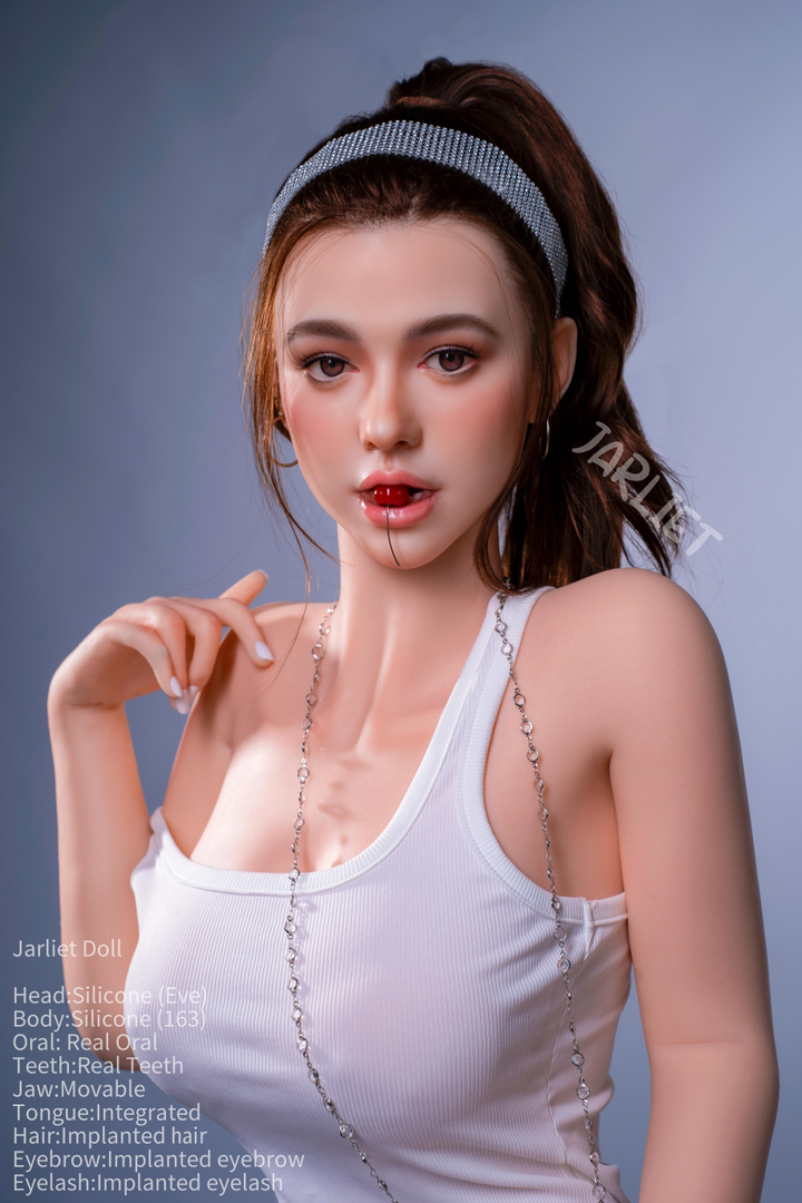 A front-facing portrait of the Jarliet Doll 163 cm D Silicone - Eve V2 featuring her movable jaw and real teeth as she holds a cherry between her lips. She is wearing a white tank top and a silver beaded headband.