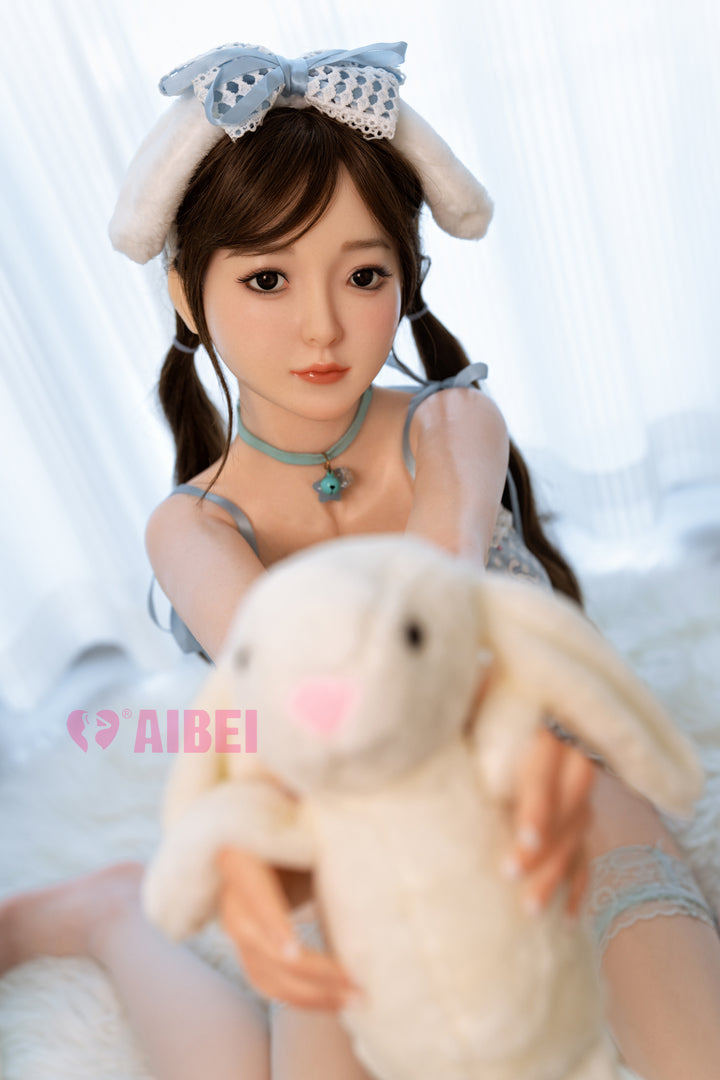 Aibei Doll 151 cm D-cup silicone model 'Yui' holding a plush bunny, styled with bunny ears headband, twin ponytails, and a delicate necklace, posed in a soft daylight setting.