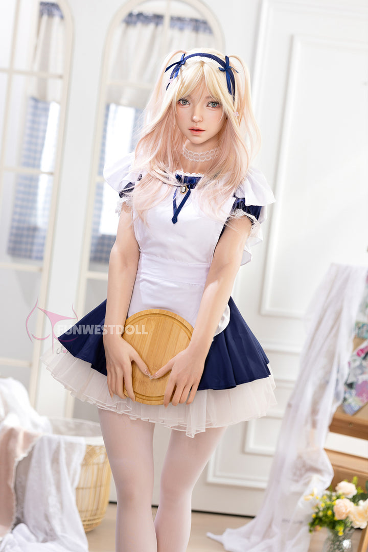 FunWest Doll 159 cm A Silicone – Giselle standing in a bright room, dressed in a blue and white maid uniform with a short skirt and apron, holding a small wooden tray.