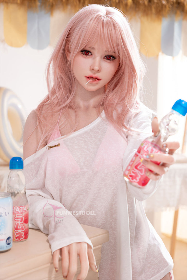 FunWest Doll 159 cm A Silicone - Lucy (Real Skin Texture) seated at a wooden table, wavy rose-pink hair with soft bangs, holding a blue water bottle, sheer white blouse slipping off one shoulder, cozy indoor setting with a blue curtain backdrop