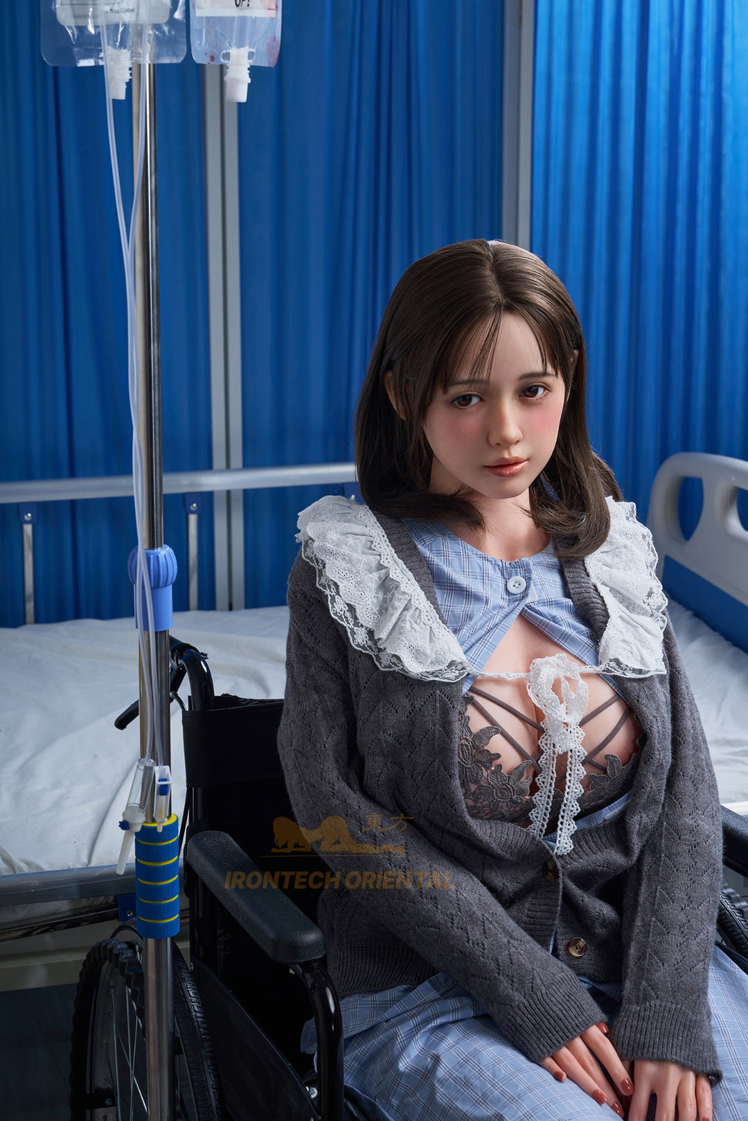 Wide shot of the Misaki V3 doll seated in a wheelchair next to an IV pole in a medical-themed room setting.
