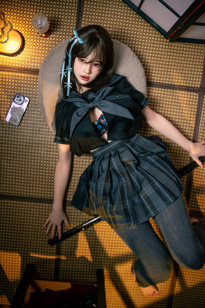 Irontech Doll 161cm F Oriental Series Silicone – Evie V4 – Seated pose, black kimono-style outfit slightly open at the collar, blue hair ribbons, dark braided hairstyle, traditional interior backdrop.