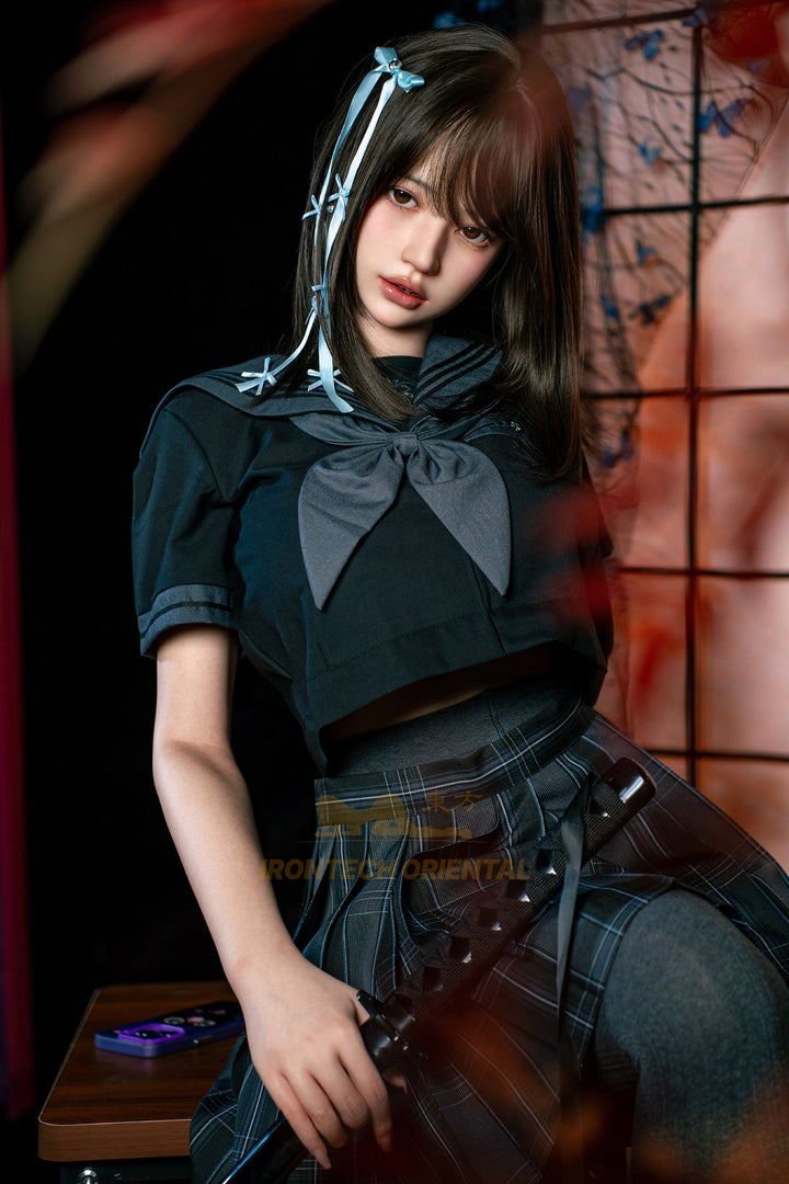 Irontech Doll 161cm F Oriental Series Silicone – Evie V4 – Seated pose leaning forward, black kimono-style top open at the chest, blue hair ribbons, dark braided hair, traditional wooden backdrop.