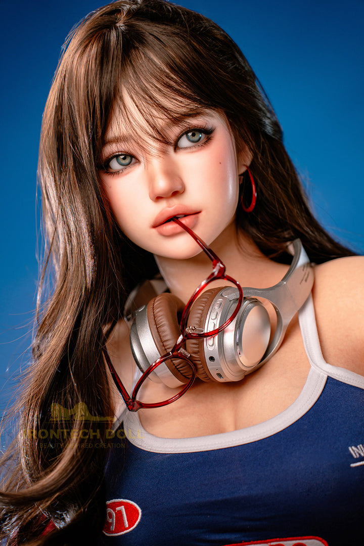 Irontech Doll 161 CM F Silicone - Evie close-up upper body view wearing red baseball cap and blue racing-style top with headphones around neck, showcasing realistic facial features with long dark brown hair against blue studio background