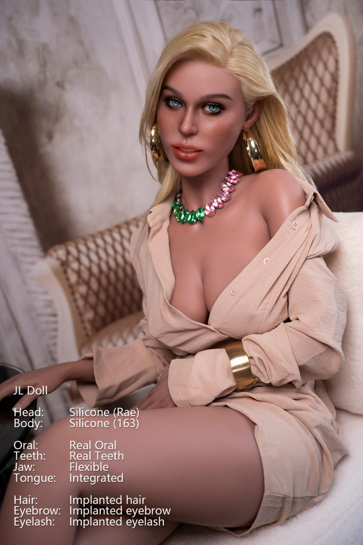 Close-up portrait of the Jarliet Doll 163 cm D Silicone - Rae with blonde hair, wearing a beige button-down shirt dress, a pink and green stone necklace, and gold jewelry while seated.
