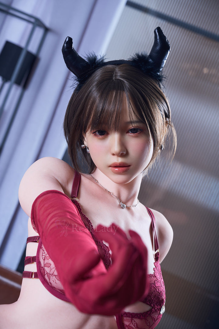 RealLady 159cm H Silicone - Alara – Close-up pose with arms raised, black horn-style headpiece, red lace bodysuit with sheer red gloves, dark bob hairstyle, softly lit interior backdrop with venetian blinds.