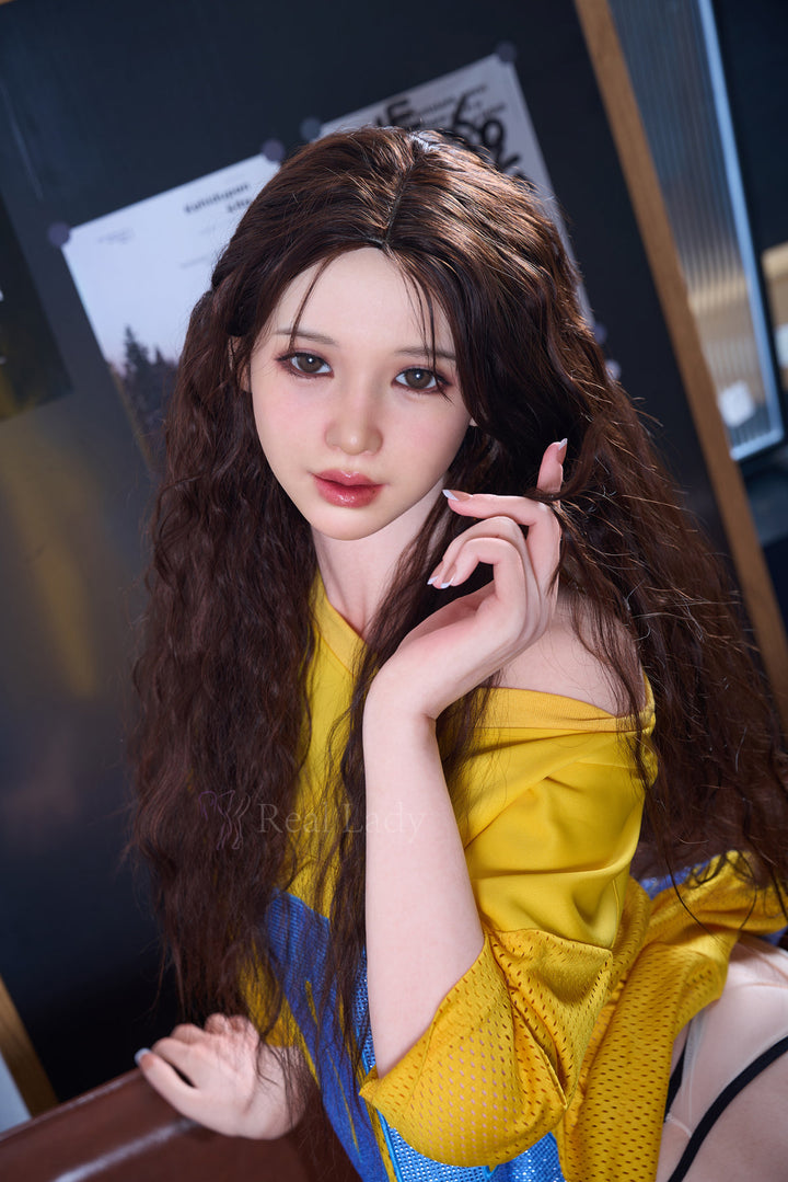 RealLady 159cm H Silicone - Alodia V3 – Close-up seated pose, hand resting near chin, long wavy dark brown hair, mustard-yellow off-shoulder dress, dark wood interior backdrop.