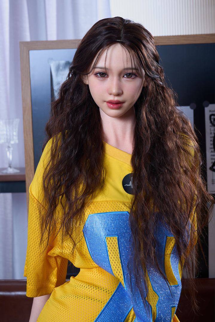 RealLady 159cm H Silicone - Alodia V3 – Seated pose with hand near face, mustard-yellow off-shoulder dress, long wavy brown hair, dark interior backdrop.