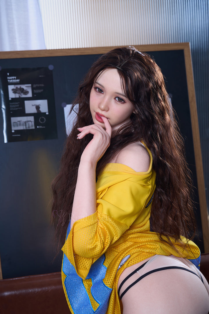 RealLady 159cm H Silicone - Alodia V3 – Seated side pose on wooden stool, mustard-yellow off-shoulder dress, long wavy brown hair, dark interior backdrop.