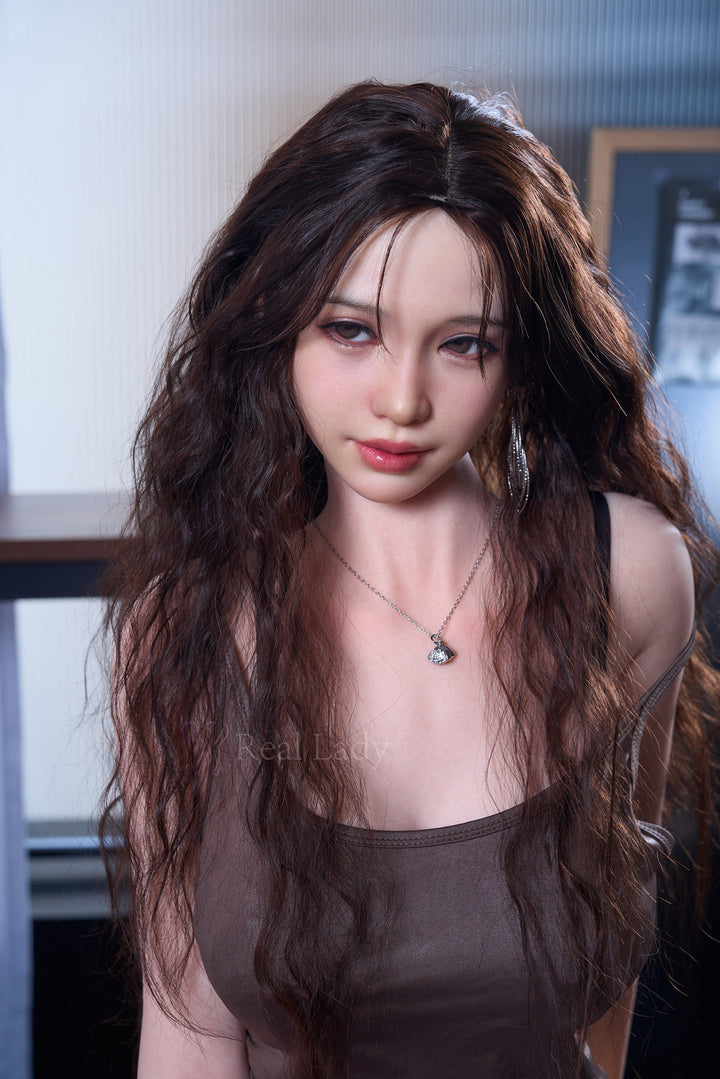 RealLady 159cm H Silicone - Alodia V4 – Seated pose leaning forward, sheer dark mesh top revealing the chest, layered jewelry, wavy brown hair falling forward, indoor backdrop.