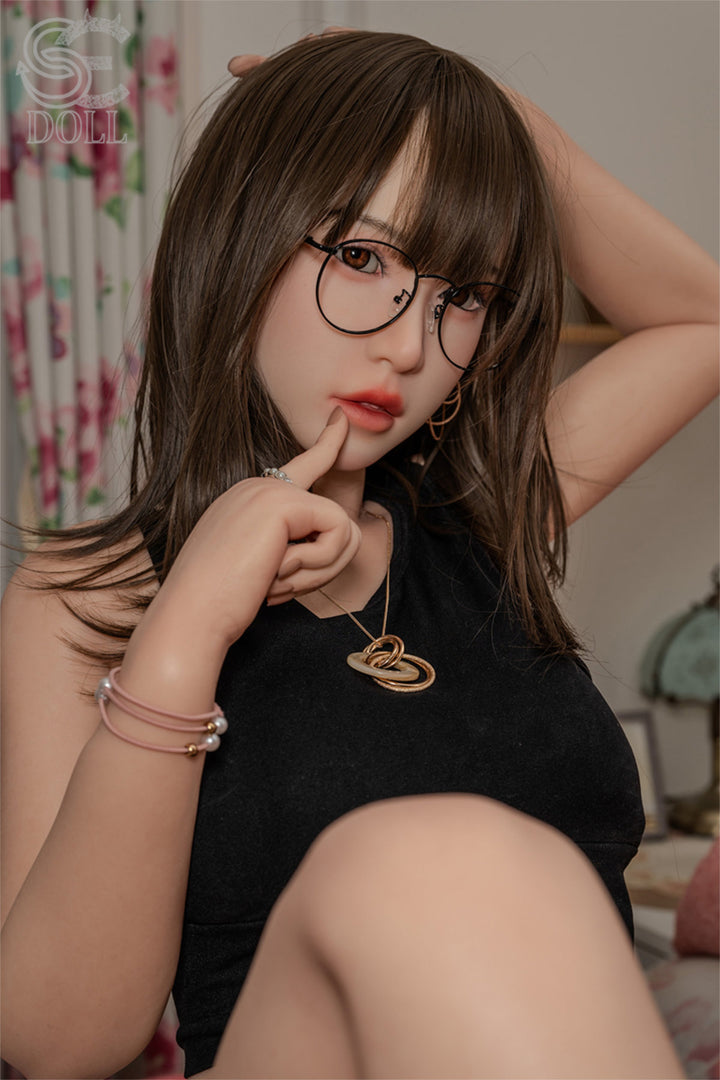 SEDOLL T153cm A Aya Series Silicone - Sena.A – Close-up pose with finger near lips, round eyeglasses, black cropped tank top, dark brown bangs and long hair, softly lit bedroom backdrop.