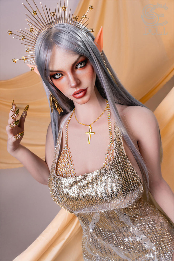 SEDOLL T167 cm E Silicone Pro - Evadora.A close-up with one hand raised near her cheek, delicate rings on her fingers, gold sequined dress strap sliding off her shoulder, warm gold silk backdrop