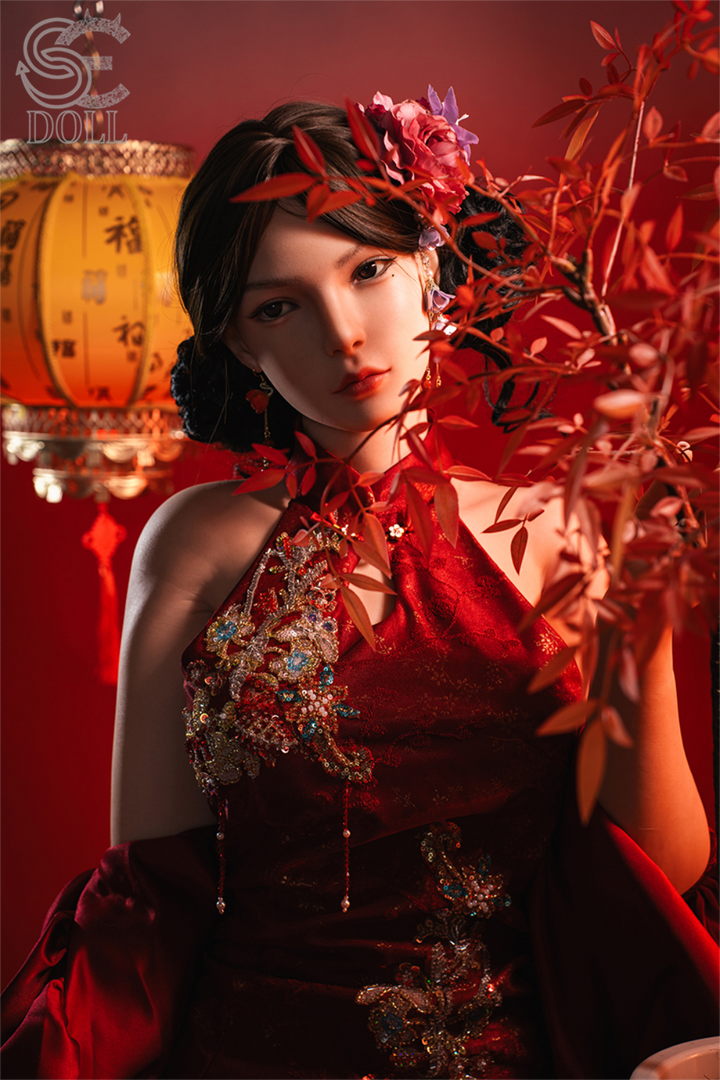 Mid-shot of the SE DOLL T165 cm C Silicone Pro - Lumi.B posing behind red foliage, dressed in a red embroidered cheongsam-style top with a traditional lantern in the background.