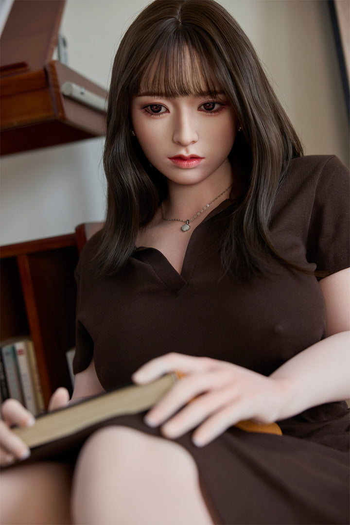 Starpery 154cm E - Yuan – Seated pose at a wooden table, brown V-neck top with buttoned front, holding a small object in one hand, dark straight-bang hairstyle, rustic wood-toned backdrop.