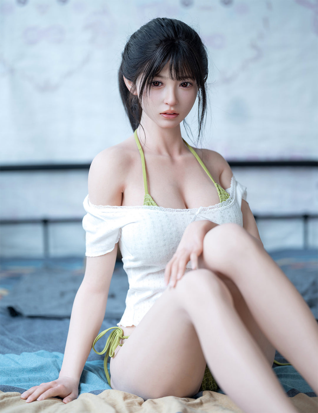 Seated front view of the Koharu doll on a bed, dressed in a casual white top and green bikini bottoms.