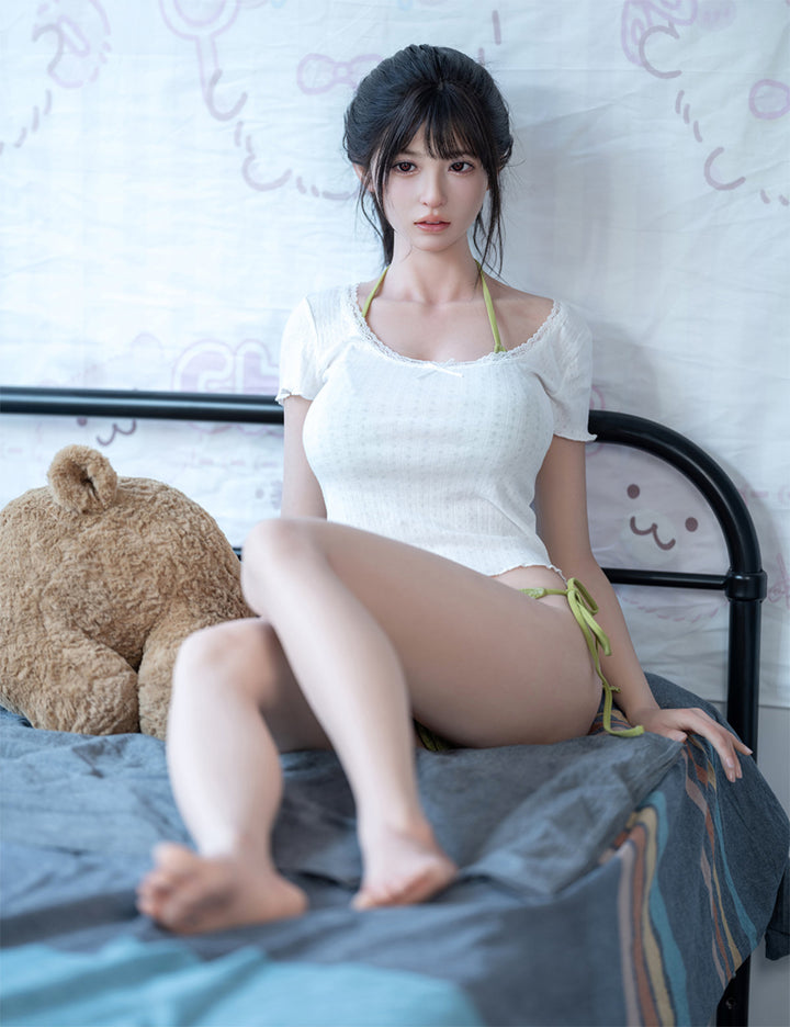 Full-length view of the Koharu doll leaning back against a black bed frame with a teddy bear in the background.