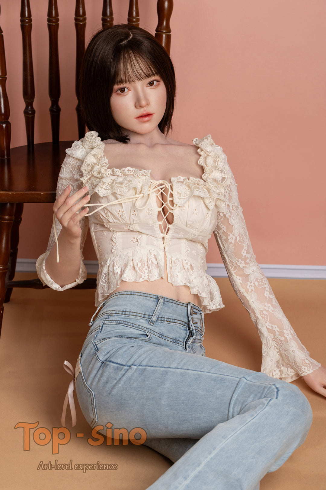 Top Sino 167 cm Platinum Silicone - Mimu (RRS+) - V2 wearing a lace top and jeans sitting on the floor in front of a brown chair against a peach wall.