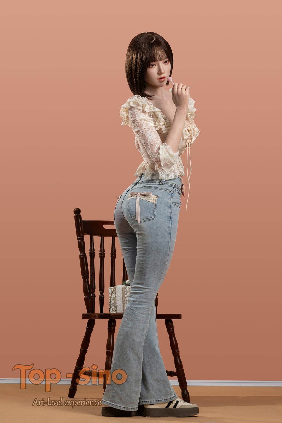 Top Sino 167 cm Platinum Silicone - Mimu (RRS+) - V2 in a white blouse and blue jeans standing in front of a wooden chair against a peach wall.