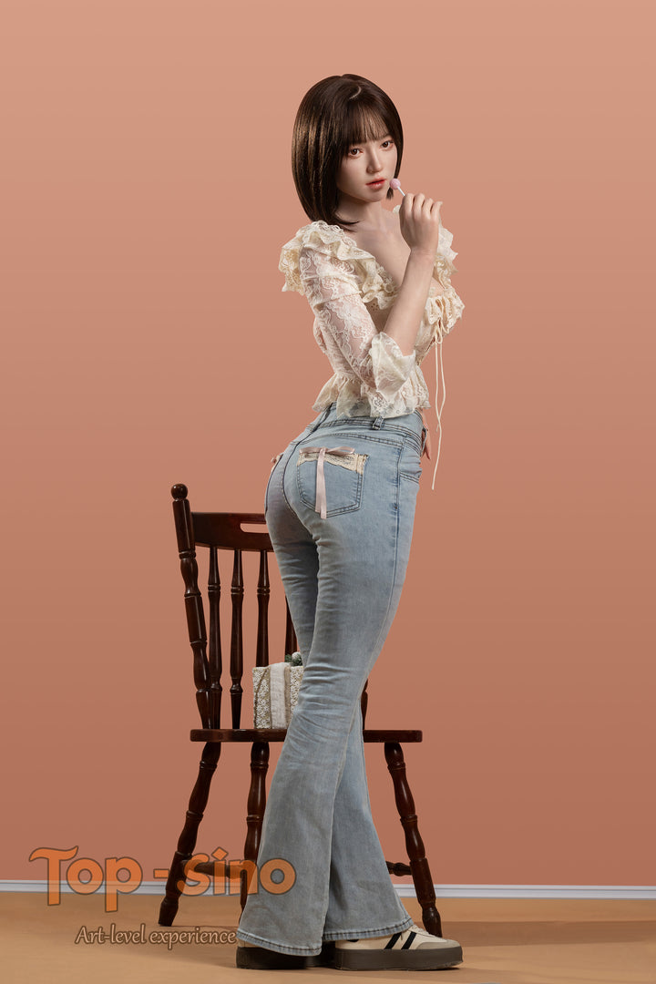 Top Sino 167 cm Platinum Silicone - Mimu (RRS+) - V2 in a white blouse and blue jeans standing in front of a wooden chair against a peach wall.