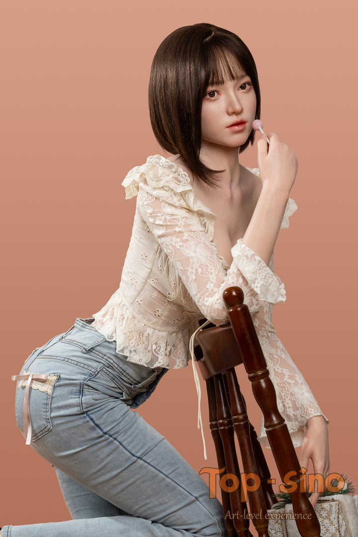 Top Sino 167 cm Platinum Silicone - Mimu (RRS+) - V2 in a beige floral lace top and blue jeans standing at the back of a chair against a peach wall.