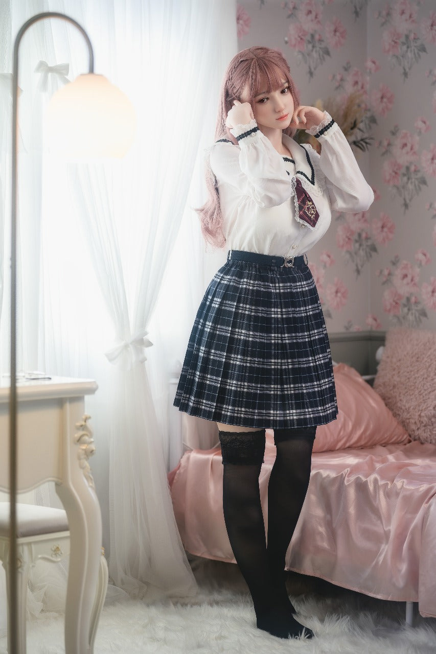 Full-body view of the doll in a schoolgirl-inspired outfit, standing by the bed with soft daylight in the background.
