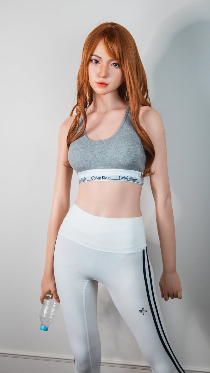Zelex Doll K-Series 168 cm B Silicone - Suyeon three-quarter standing view in grey sports bra and white athletic leggings, long auburn hair, white studio background
